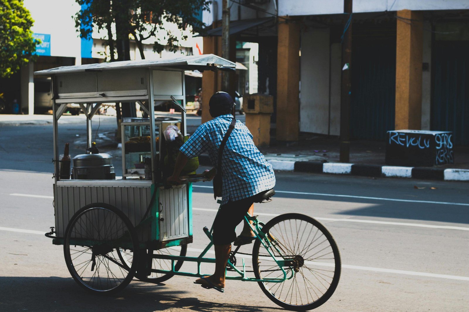 An adult male vendor rides a bicycle food cart through a city street on a sunny day